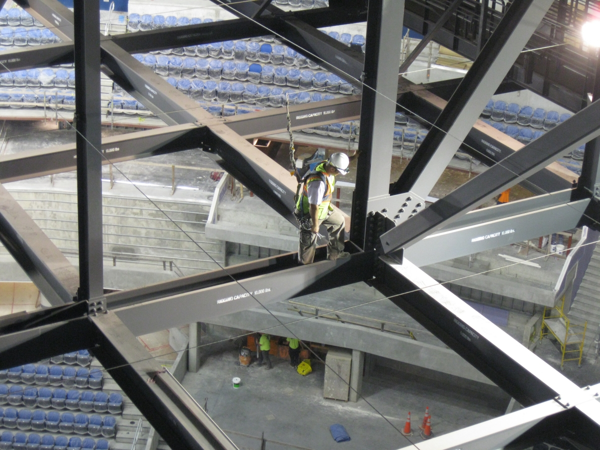 Worker in safety harness navigating structural beams in an arena under construction, highlighting fall protection measures for worker safety in high-risk environments.